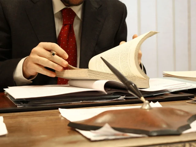 En primer plano una pluma, en segundo las. manos de un abogado en su escritorio leyendo la Ley Federal de Procedimiento Contencioso Administrativo.